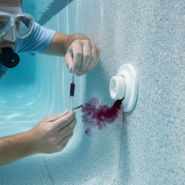 Underwater pool leak detection dye test in progress, technician wearing a light blue t-shirt using a  small syringe to release small amount of dark red dye around a pool fitting or patch, dye visibly drawn into a small opening indicating a leak, clear pool water, textured pool surface visible, close-up professional diagnostic process, non-invasive inspection method, r
